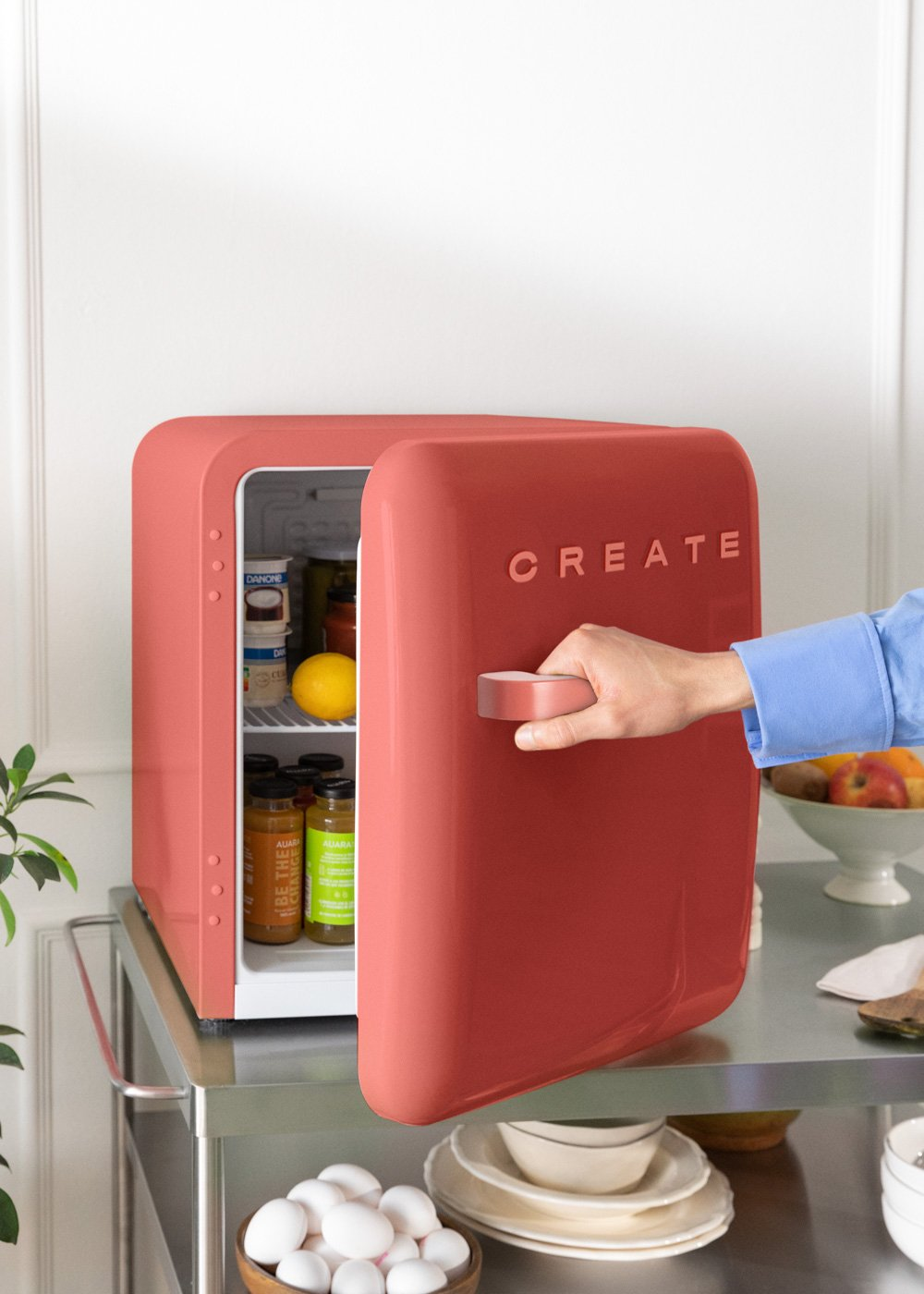 Mini nevera coral con la puerta abierta. Mano abriendo la puerta. Dentro hay tarros, fruta y otros artículos.
