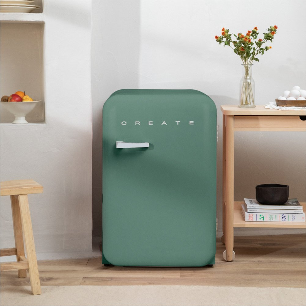 Un refrigerador verde con la palabra CREATE. Flores a la derecha. Un taburete y fruta a la izquierda.