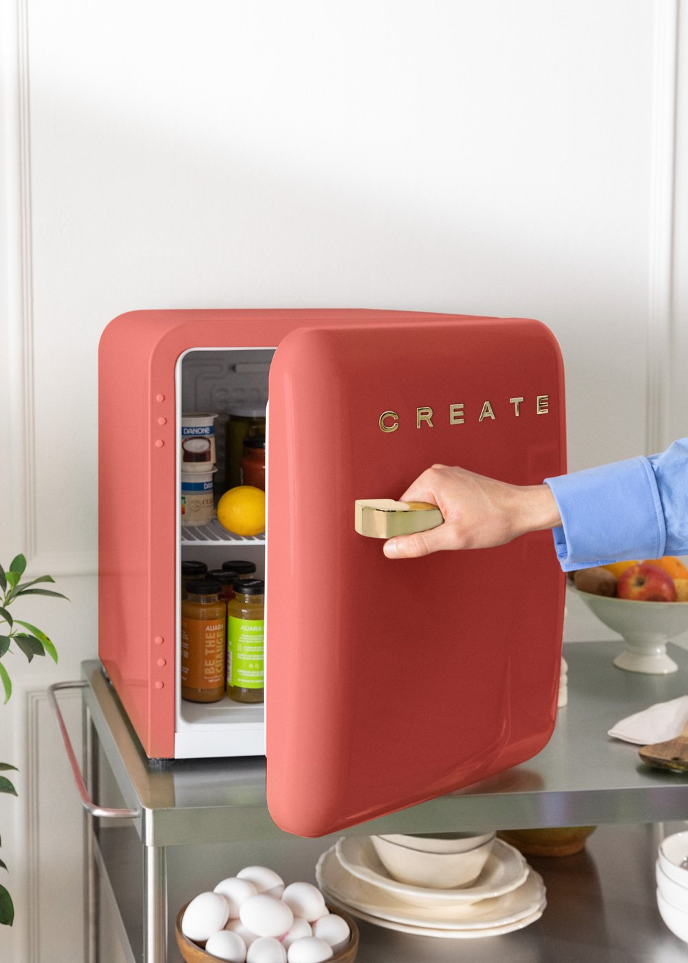 Una mano abre un refrigerador coral con CREATE. El refrigerador está en un carrito con comida dentro.