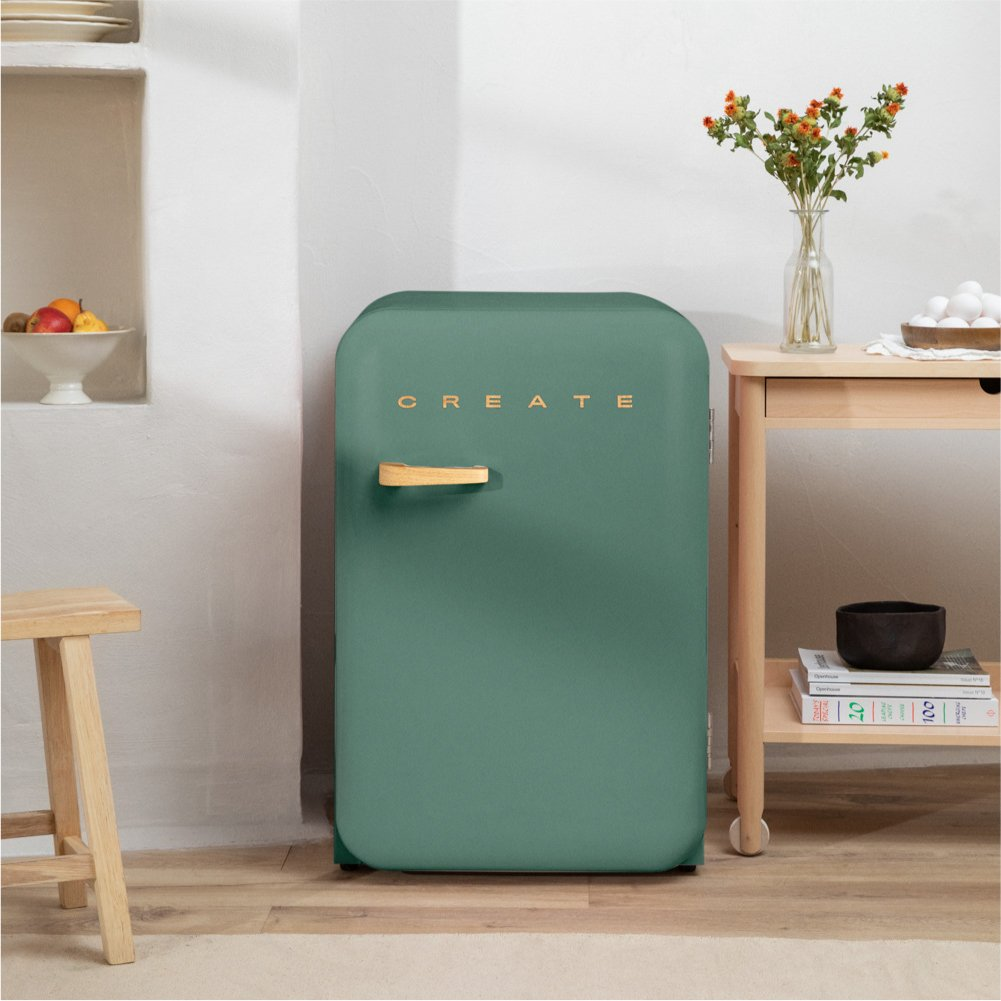 Un mini refrigerador verde con un asa de madera se encuentra en un piso de madera junto a una mesa pequeña.
