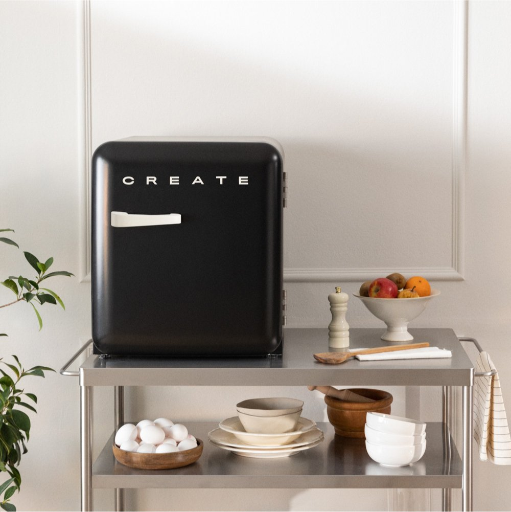 Un mini refrigerador negro se encuentra en un carrito de metal, rodeado de utensilios de cocina y frutas.