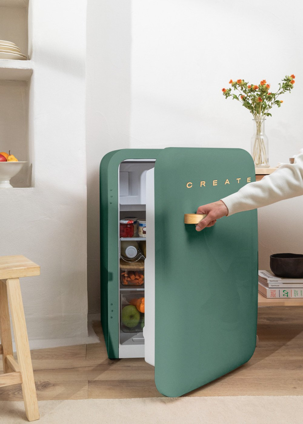 Un mini refrigerador verde abierto revela alimentos en su interior, junto a un taburete y una mesa de madera.