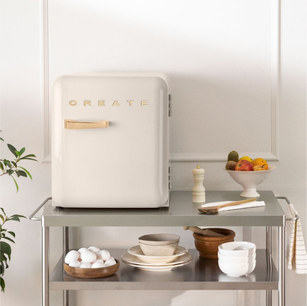 Un mini refrigerador blanquecino se encuentra en un carrito de metal, rodeado de utensilios de cocina y frutas.