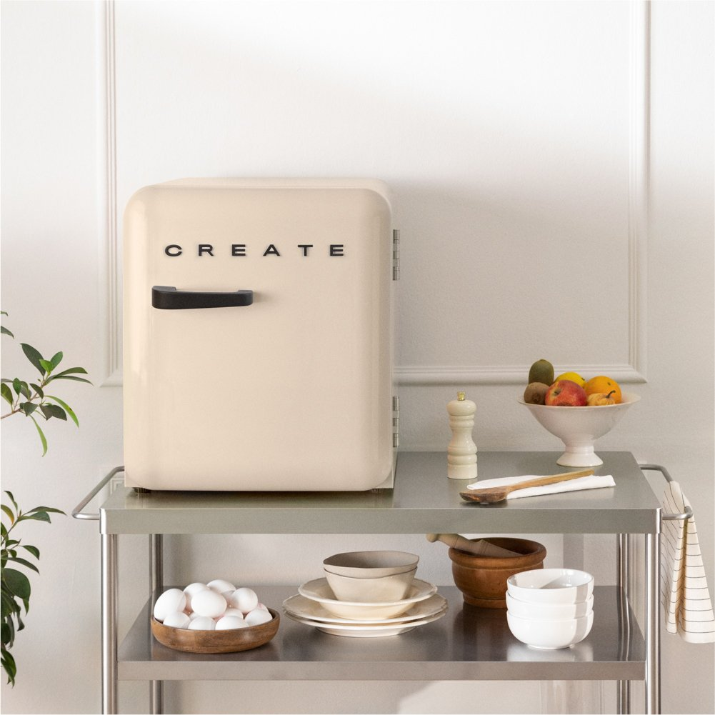 Un mini refrigerador de color crema se encuentra en un carrito de metal, rodeado de utensilios de cocina y frutas.
