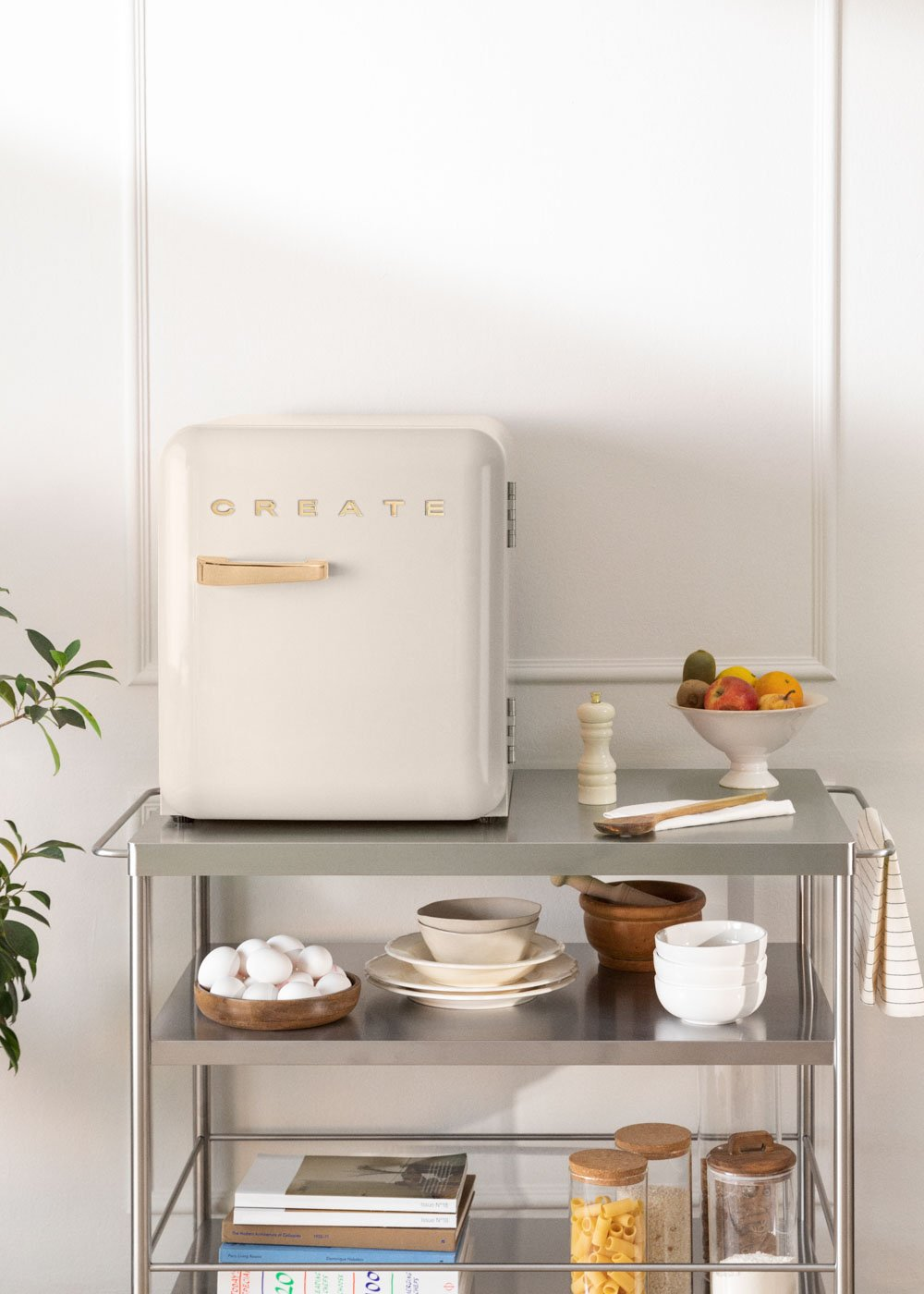 Un mini refrigerador de color crema se encuentra en un carrito de metal, rodeado de utensilios de cocina y frutas.