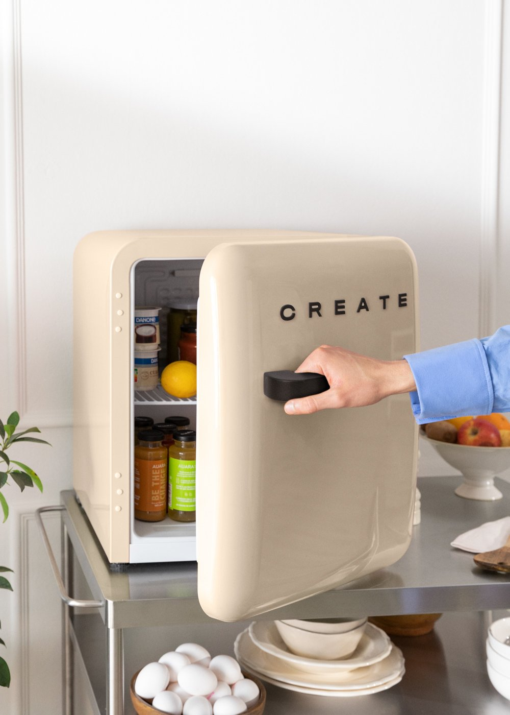 Un mini refrigerador crema abierto muestra comida en su interior, con una mano en la manija negra de la puerta.
