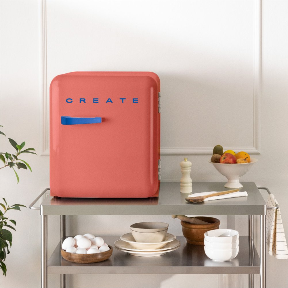 Un mini refrigerador de coral con un asa azul se encuentra en un carrito de metal, rodeado de frutas y utensilios de cocina.