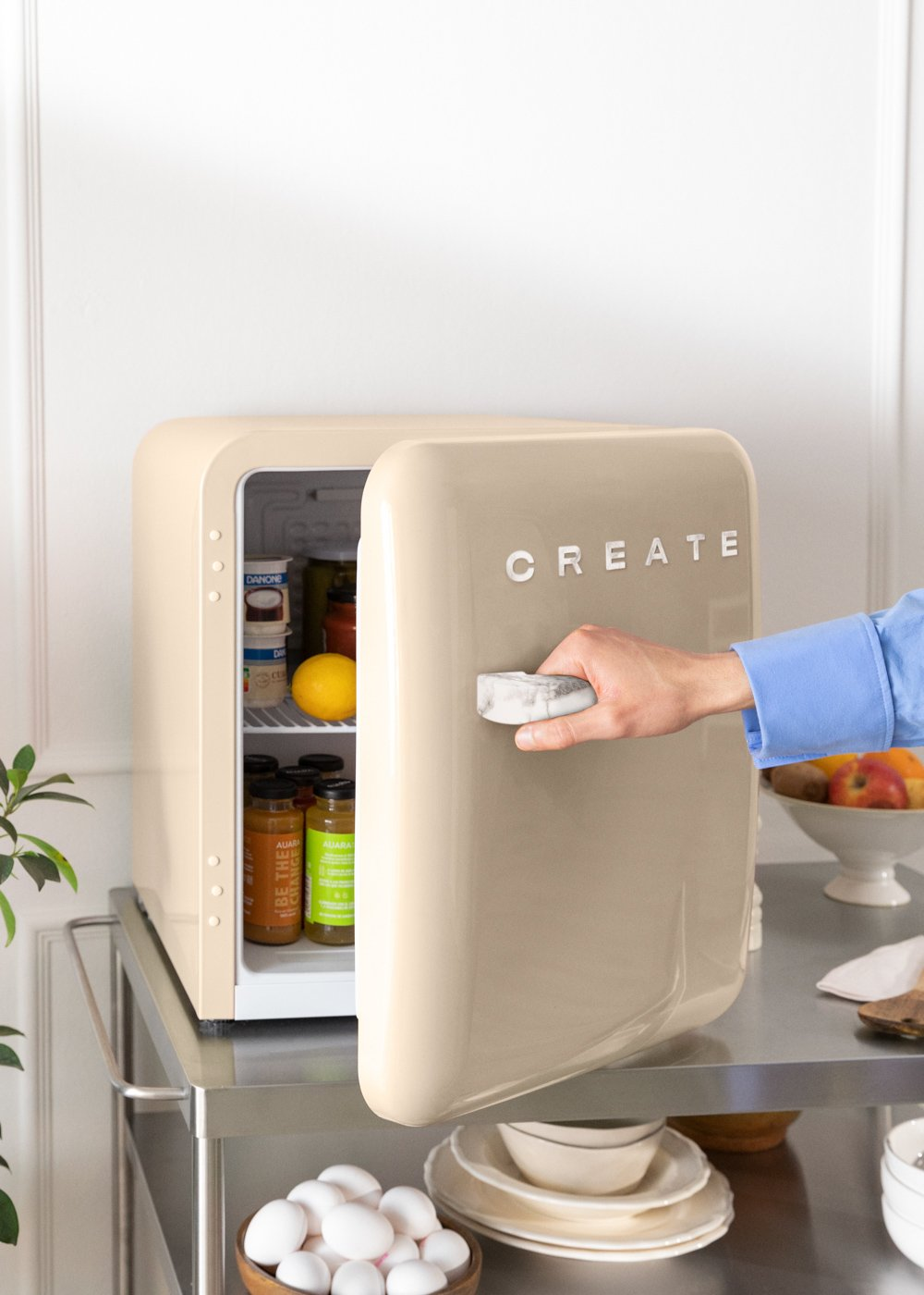 Un pequeño refrigerador crema está abierto. Una mano sostiene el tirador. Hay comida dentro.