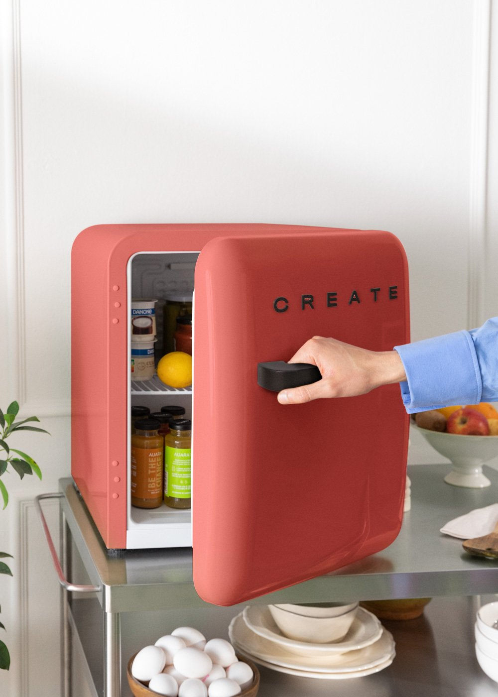 Una mano abre un mini-refrigerador coral con CREATE, lleno de artículos en su interior.