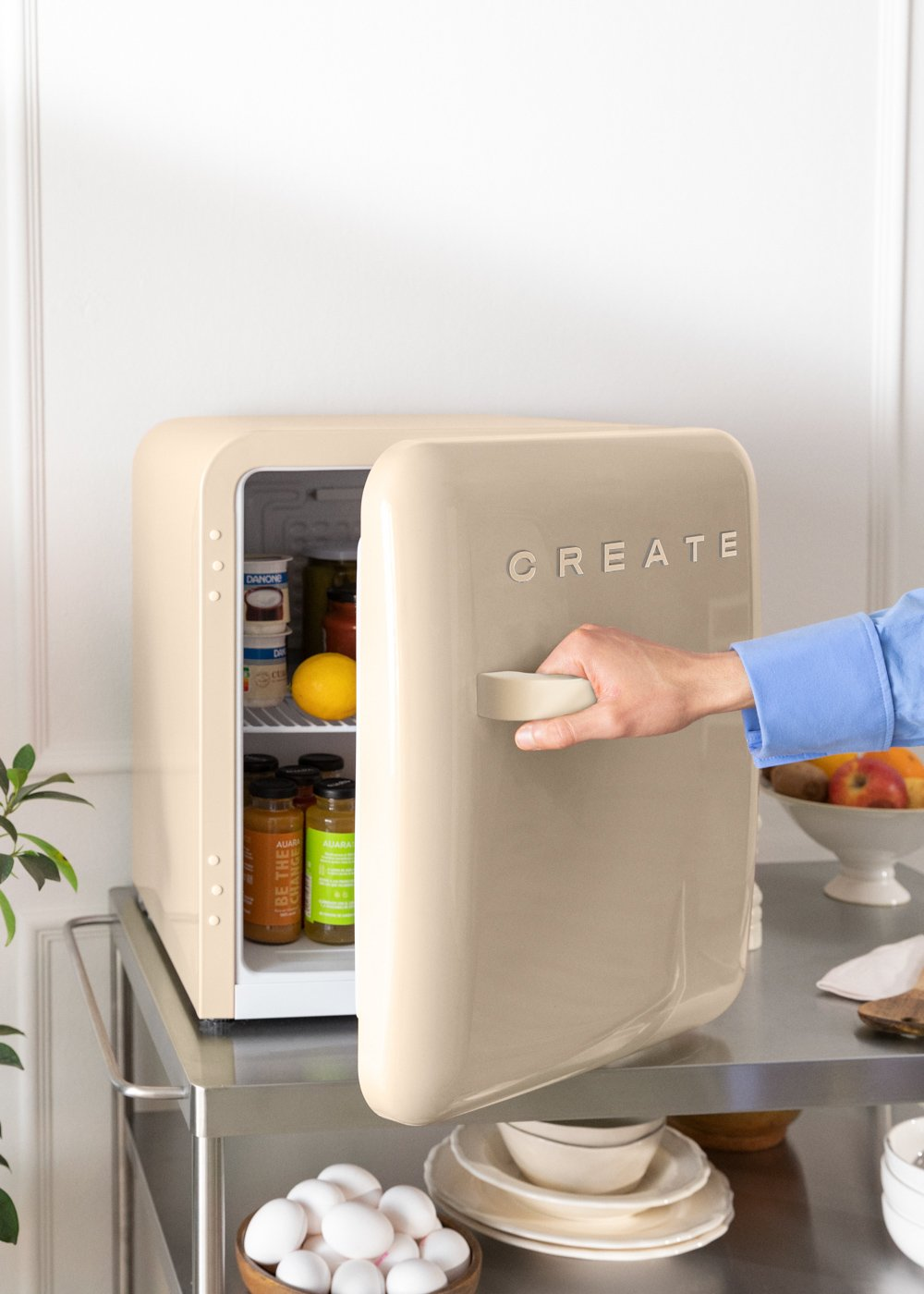 Una mano abre un mini-refrigerador beige con CREATE, lleno de artículos en su interior.