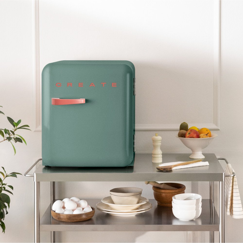 Un mini-refrigerador turquesa con CREATE sobre un carrito de metal con huevos y fruta.