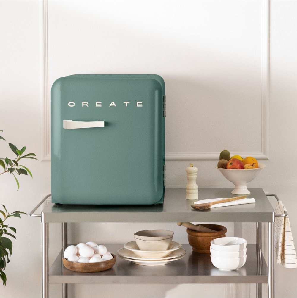 Una mini nevera verde con 'CREATE' en blanco está en un carrito. Cuencos de fruta, huevos y platos están al lado.