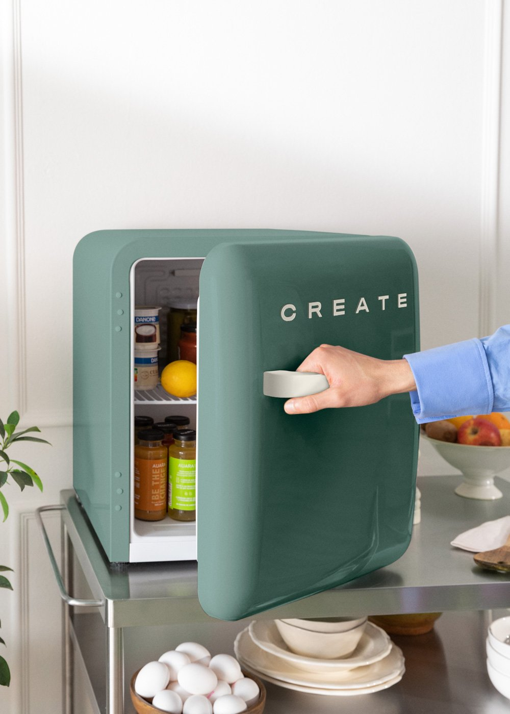 Una mano abre una mini nevera verde con la palabra 'CREATE' en blanco. En el interior, se ven alimentos. La nevera está en un carrito.
