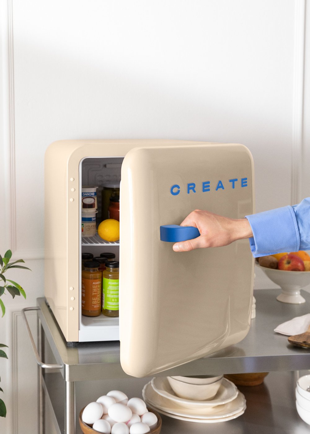 Una mano abre una mini nevera beige con la palabra 'CREATE' en azul. En el interior, se ven alimentos. La nevera está en un carrito.