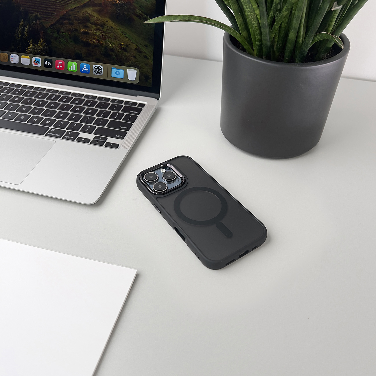 Czarne etui na telefon z laptopem, rośliną i papierem na białym biurku. Etui jest na biurku.