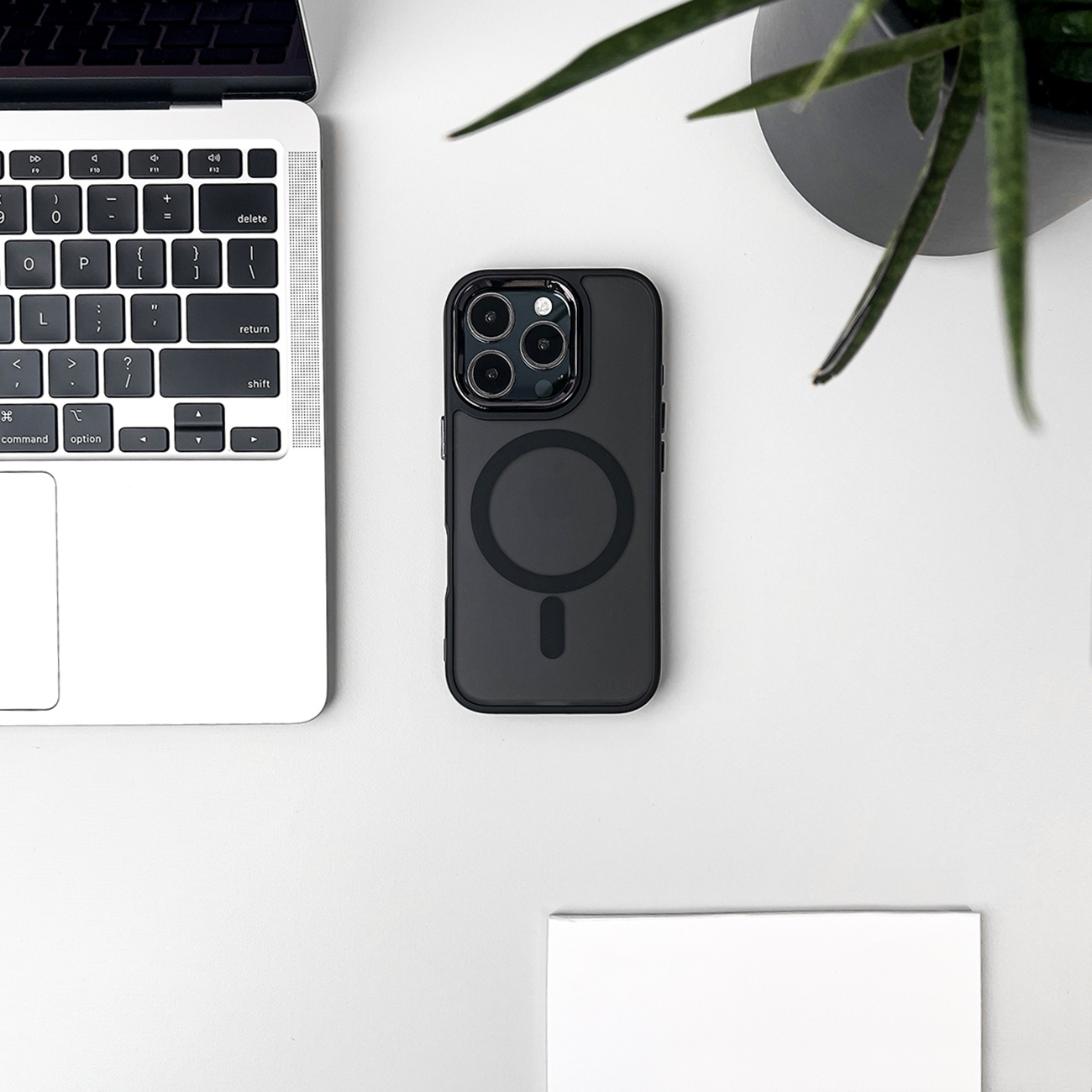 Czarne etui na telefon w pobliżu laptopa, rośliny i papieru na białym biurku. Etui jest na biurku.