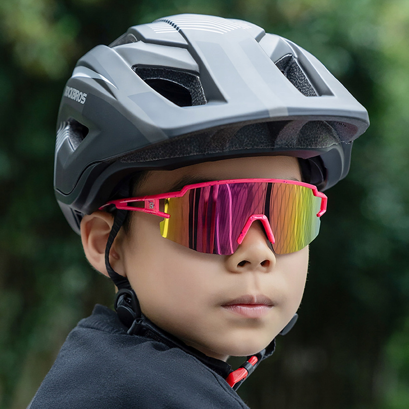 Ein Kind trägt einen grauen Rockbros-Helm und eine pink-gelbe Sportsonnenbrille. Das Kind schaut in die Kamera.