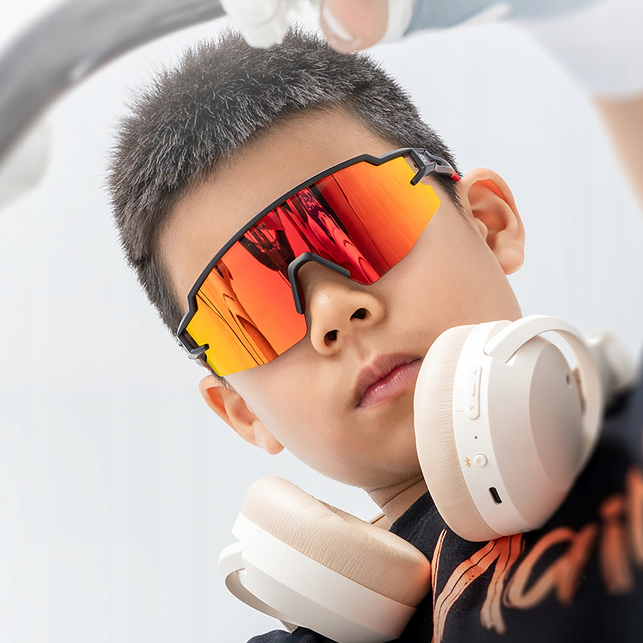 Ein Junge mit Sportsonnenbrille mit rot-orangefarbenen Gläsern. Weiße Kopfhörer um seinen Hals.