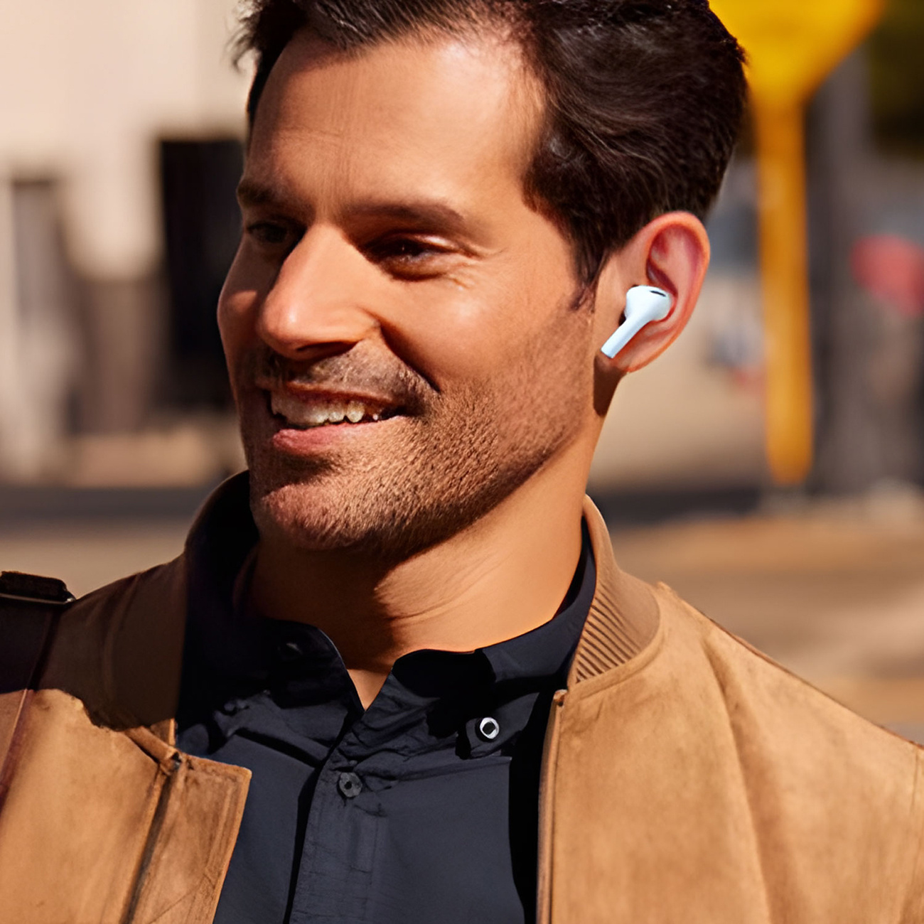 Mężczyzna ze słuchawką w uchu, uśmiechnięty, w kurtce, spacerujący na zewnątrz w słoneczny dzień.