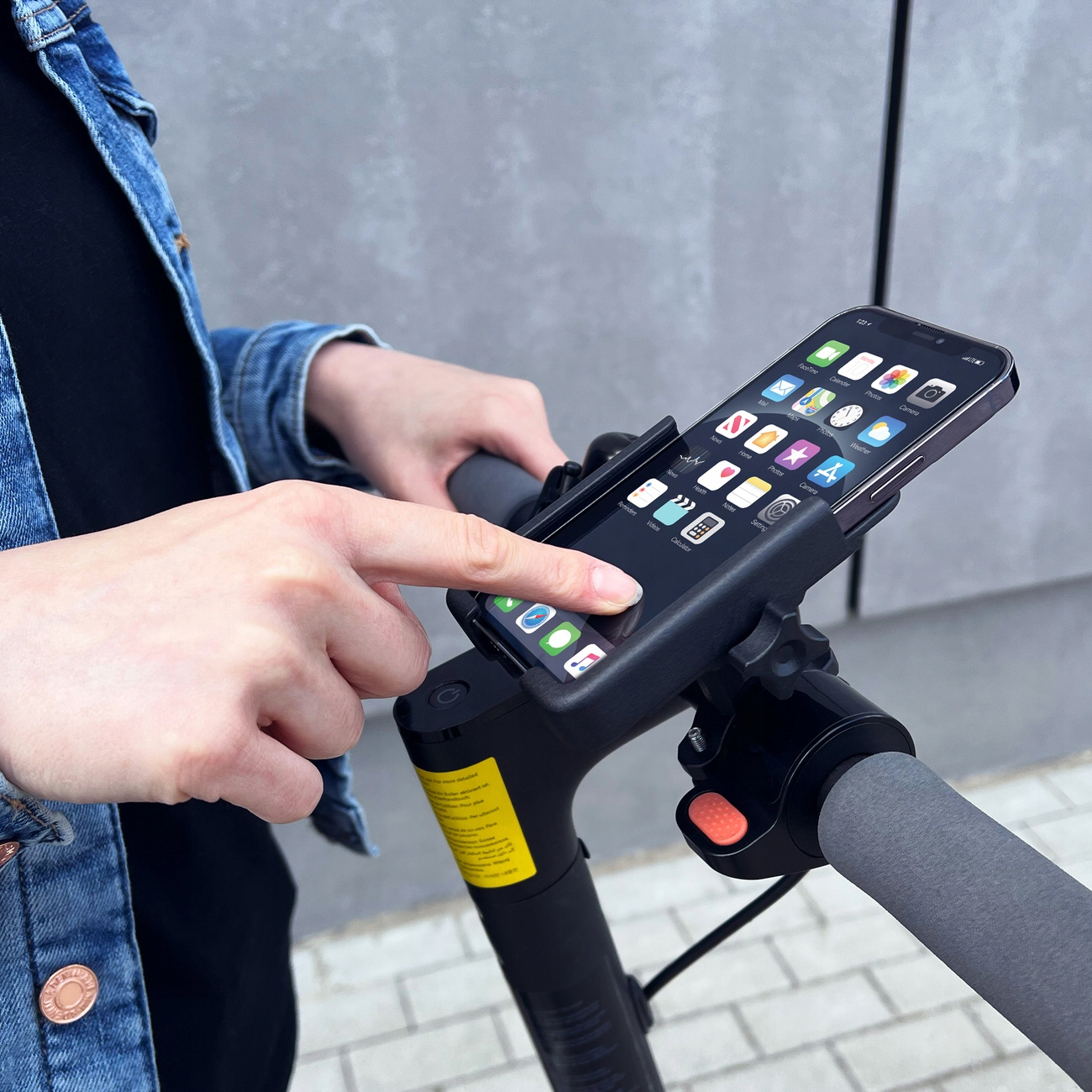 Die Hand einer Person berührt ein Telefon auf einem Elektroroller. Das Telefon zeigt Apps an. Jeansjacke und graue Lenkergriffe sind sichtbar.