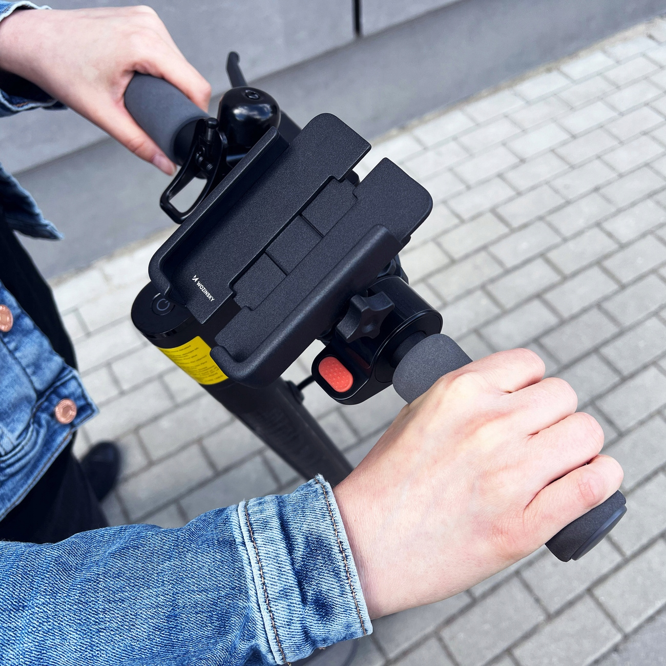 Hände halten einen Elektroroller-Lenker mit Telefonhalter. Roter Knopf und gelber Aufkleber sind ebenfalls sichtbar.