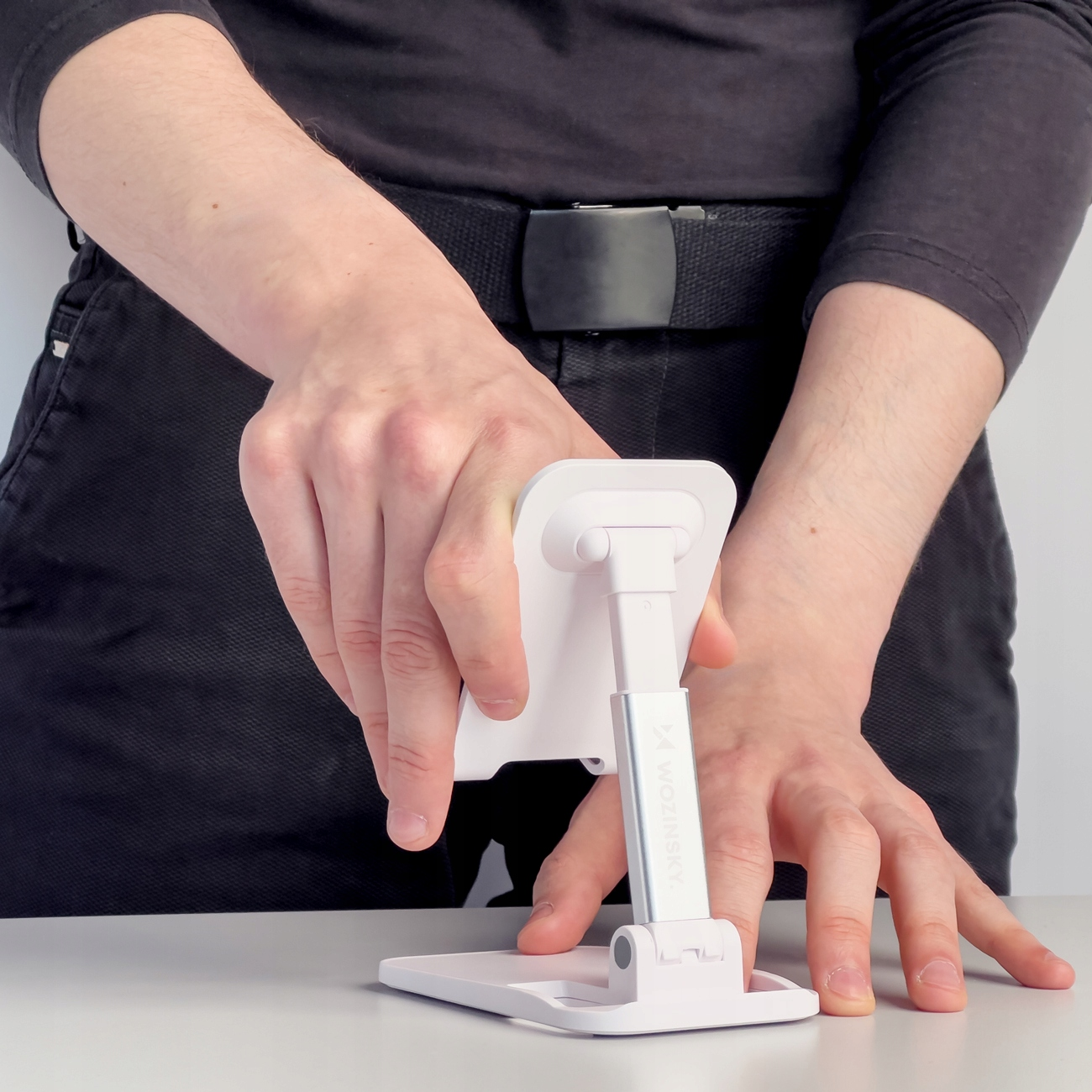 Osoba ustawiająca biały stojak na telefon na stole, z widocznymi dłońmi.