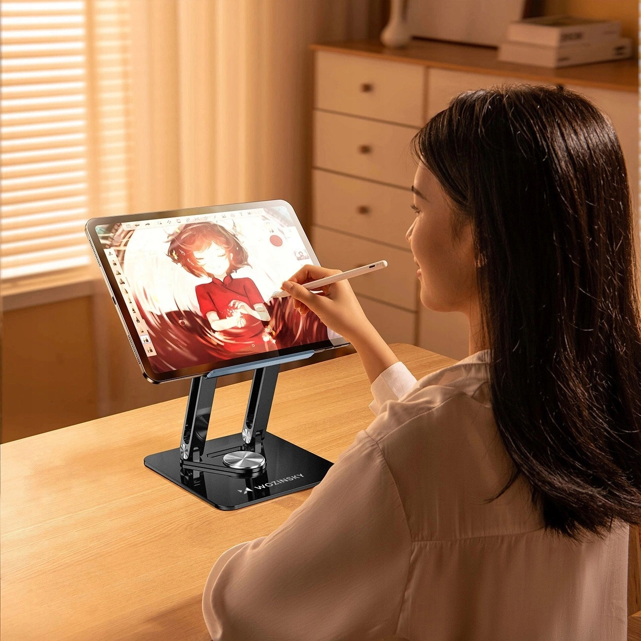Kobieta rysująca na tablecie na stojaku. Stół ma drewnianą powierzchnię. Czarny stojak.