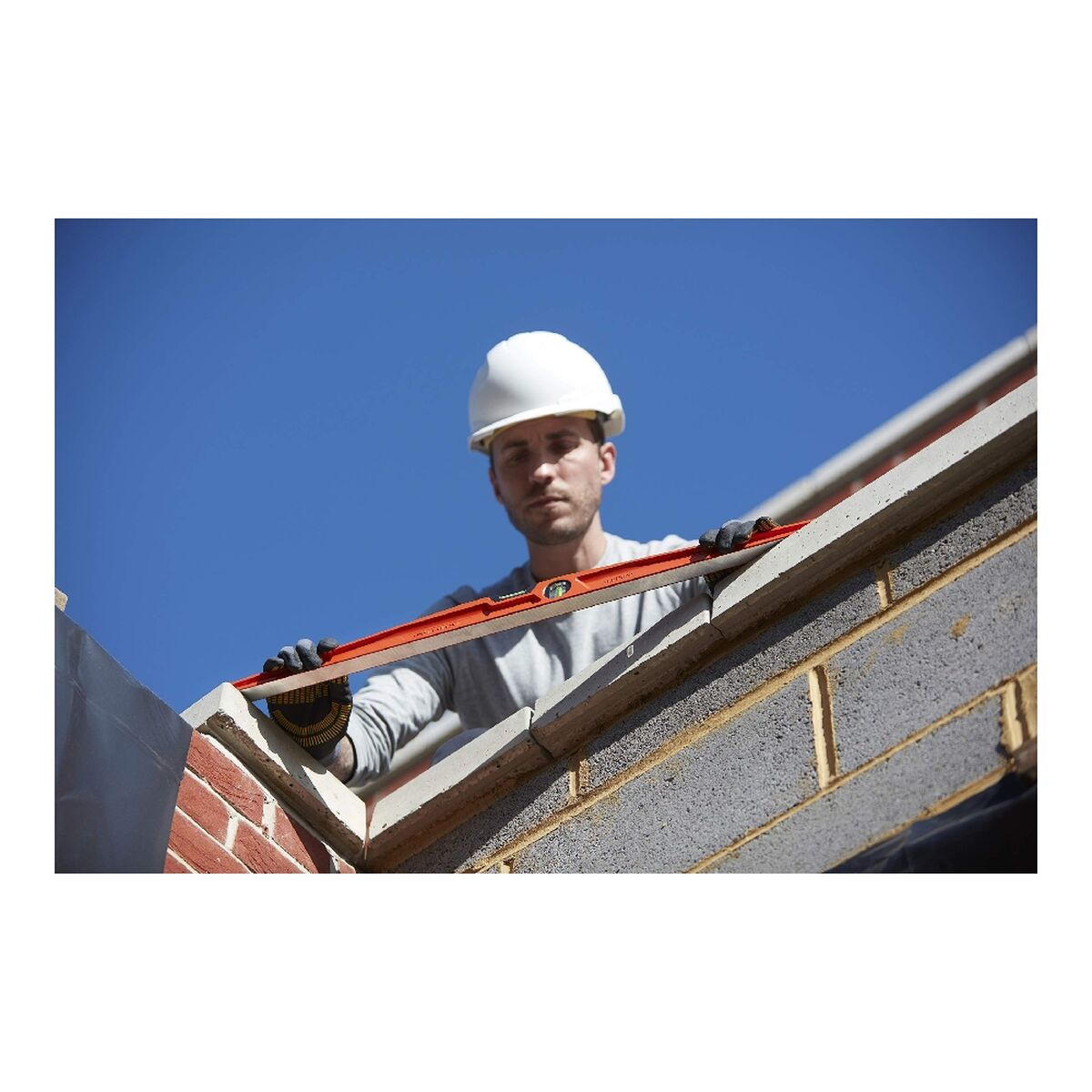 Un trabajador de la construcci ón con casco usa un nivel en una estructura de ladrillo contra un cielo azul.