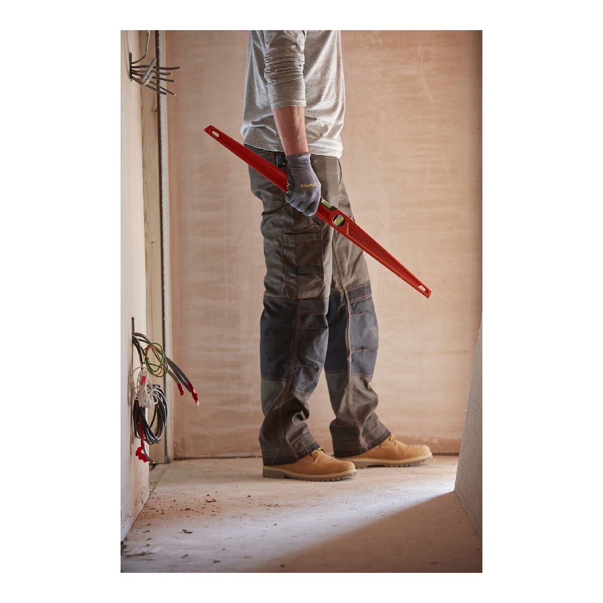 Un hombre con ropa de trabajo sostiene un nivel rojo. El fondo es una habitación sin terminar.