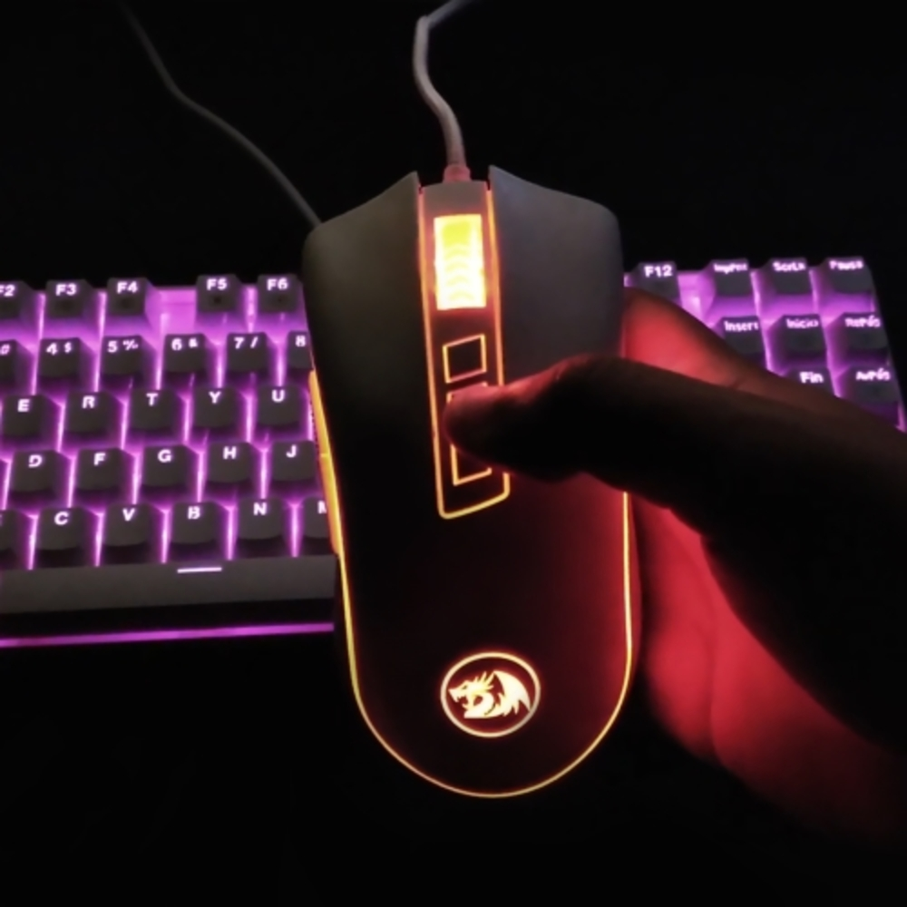 Ręka trzyma białą mysz gamingową. Podświetlenie RGB i klawiatura w tle.