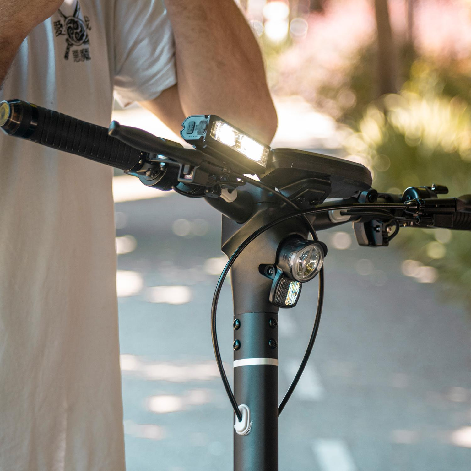 Persona sosteniendo manillar de patinete con luces, escena soleada al aire libre.