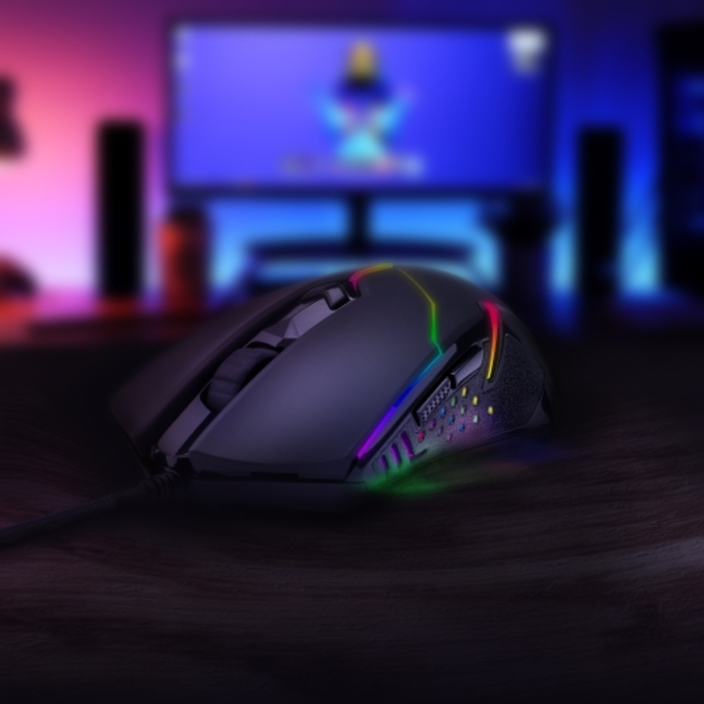 Mouse da gioco nero con luci RGB su superficie scura, TV e altoparlanti sullo sfondo.