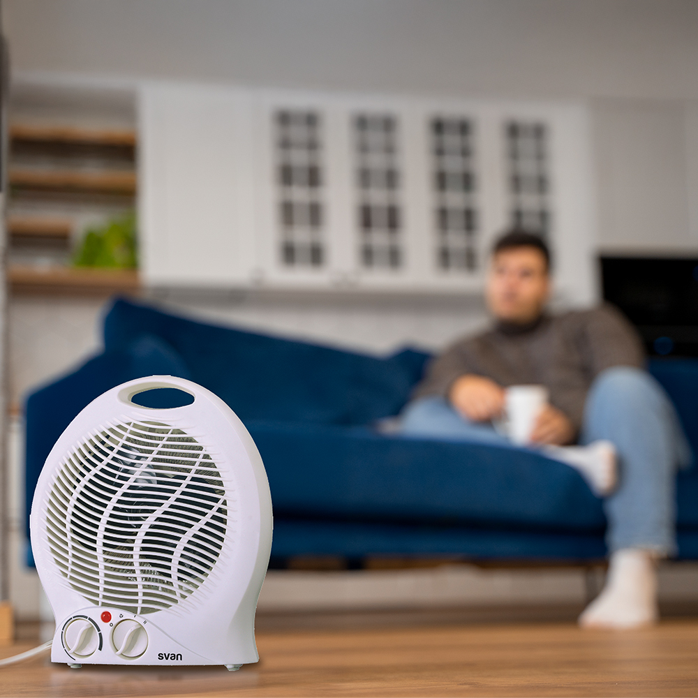 Calentador con ventilador blanco en el suelo. Un hombre está sentado en un sofá azul en el fondo, con una taza.
