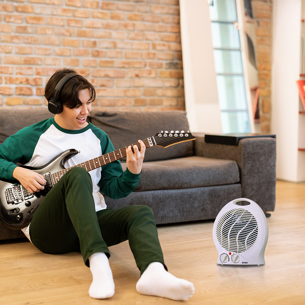 Adolescente tocando la guitarra junto a un calefactor con ventilador blanco. Sentado en el suelo, con auriculares.