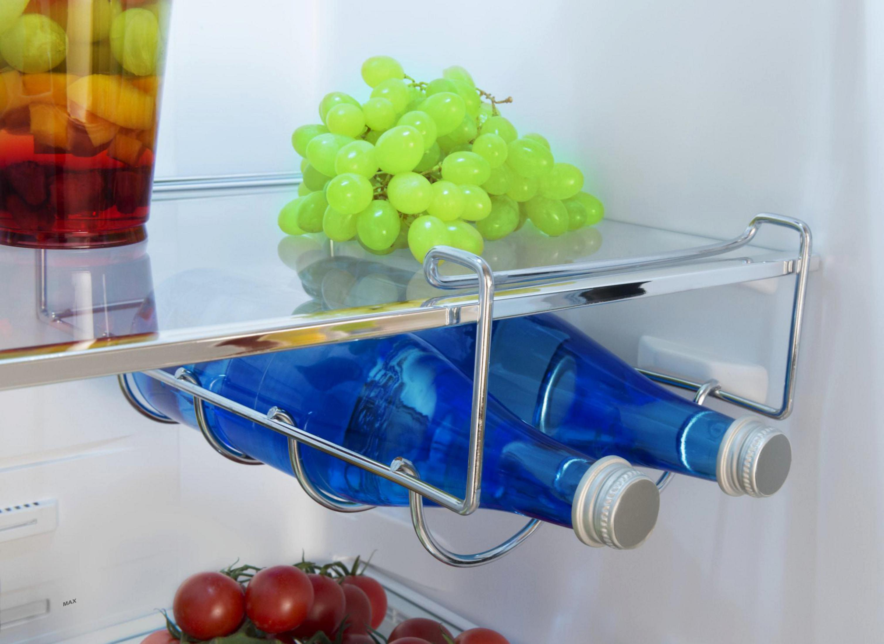 Ein Kühlschrankregal mit einem Becher Obst, Trauben, Tomaten und zwei blauen Flaschen in einem Metallgestell.