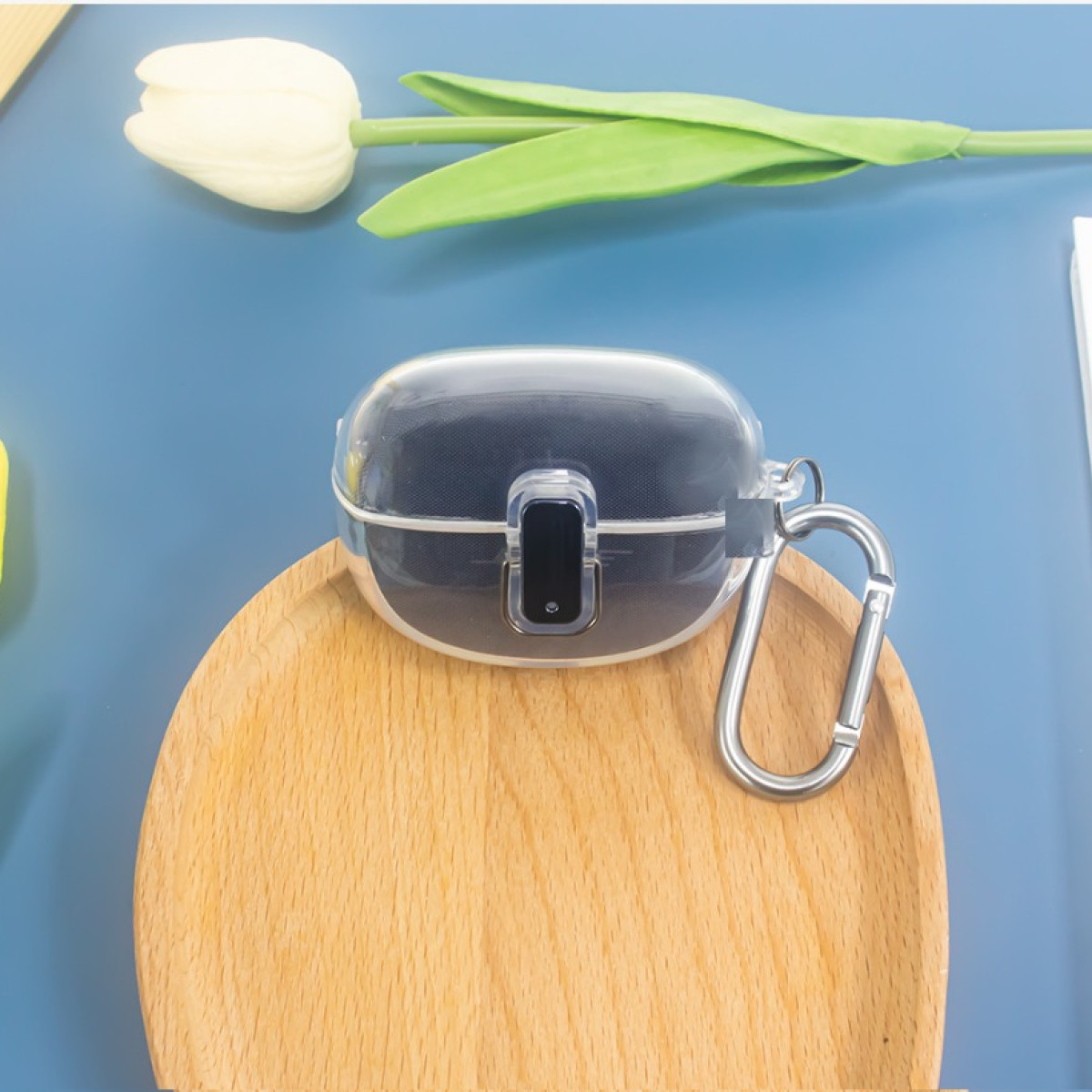 Una custodia trasparente per auricolari è su un vassoio di legno. Un tulipano bianco è in alto a sinistra. Una clip argentata fissa la custodia.