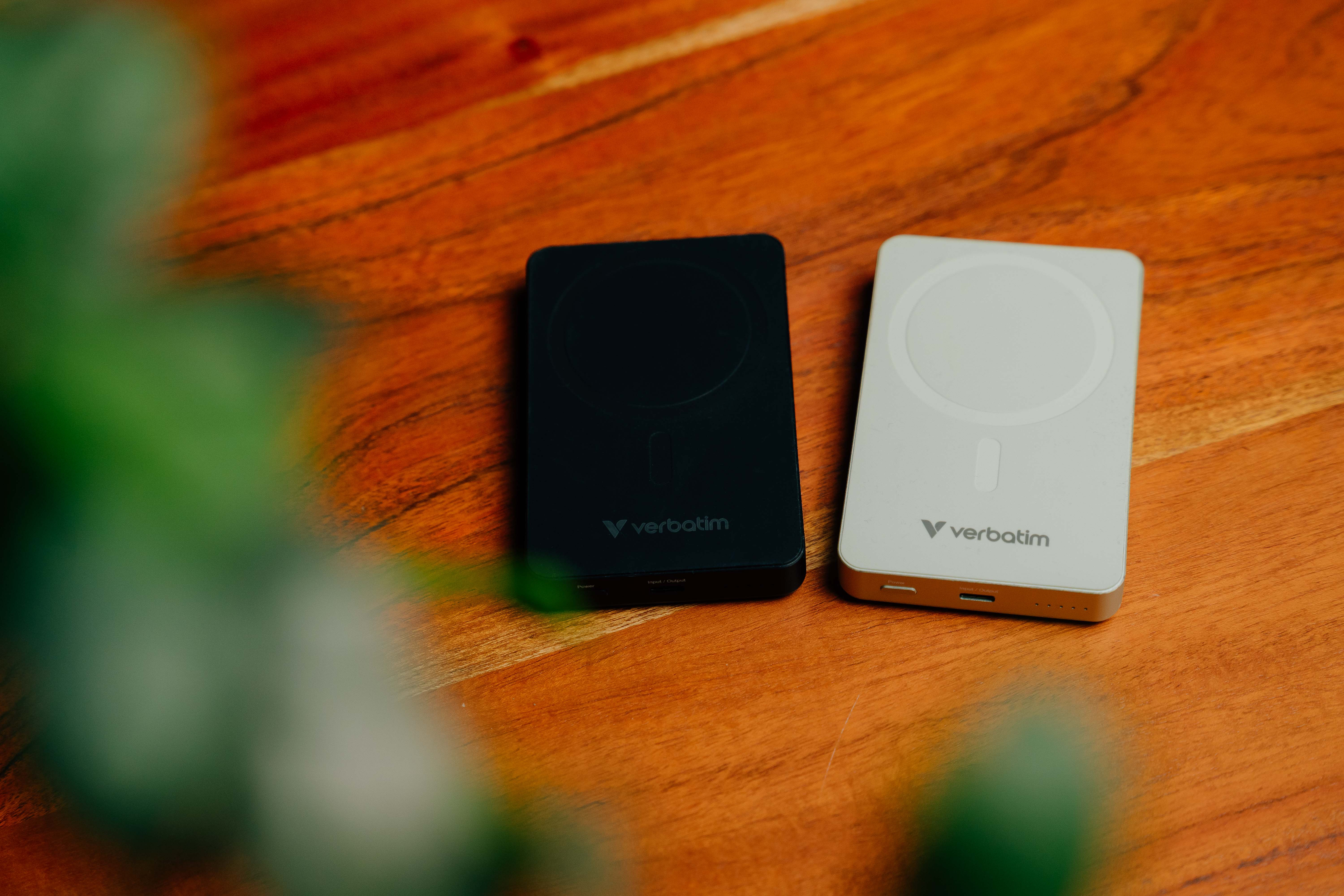 Zwei Verbatim Powerbanks auf einer Holzoberfläche, eine schwarz und eine weiß, mit Laub im Vordergrund.