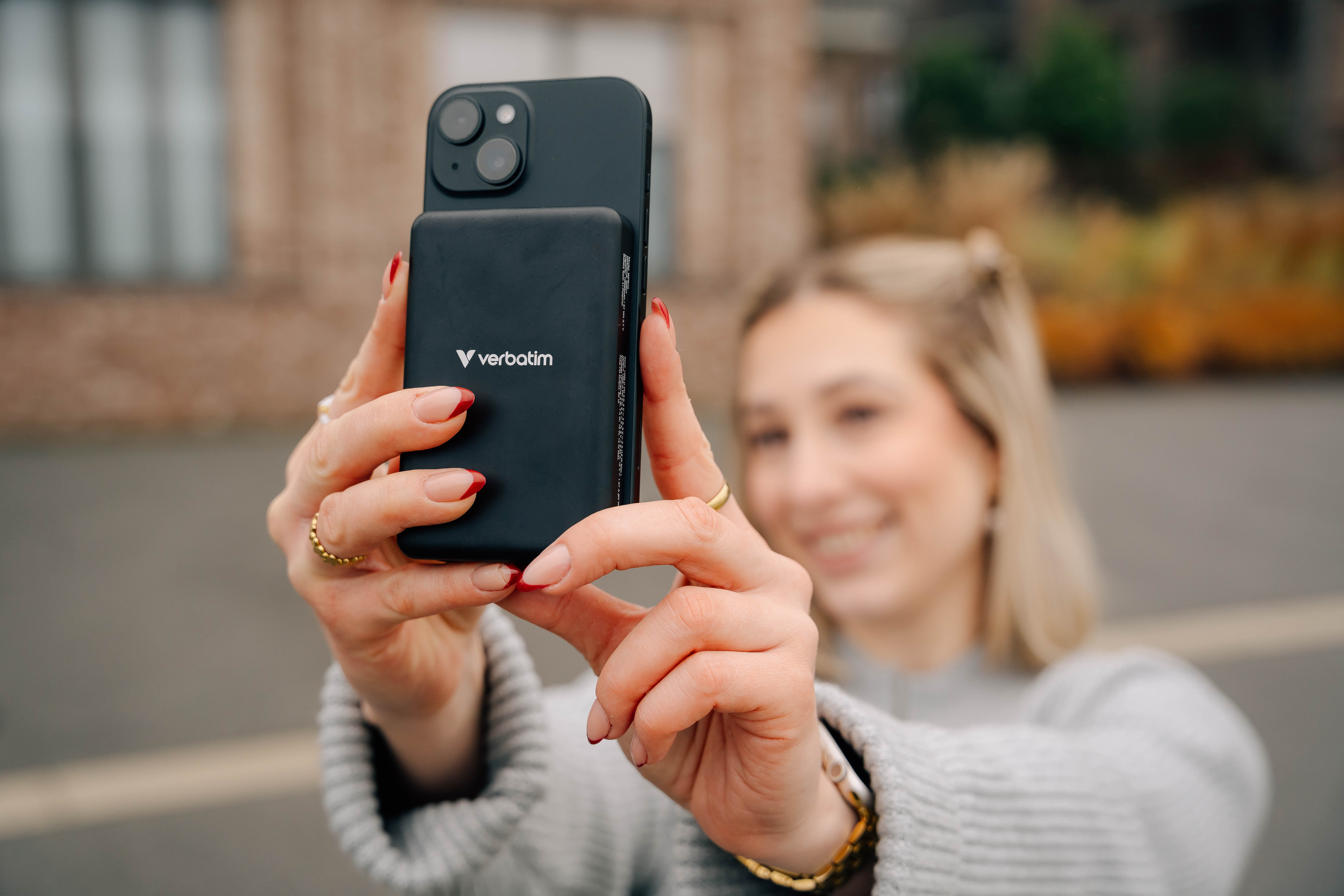 Eine Frau hält eine schwarze Powerbank und ein Telefon und macht ein Selfie. Der Hintergrund ist verschwommen.