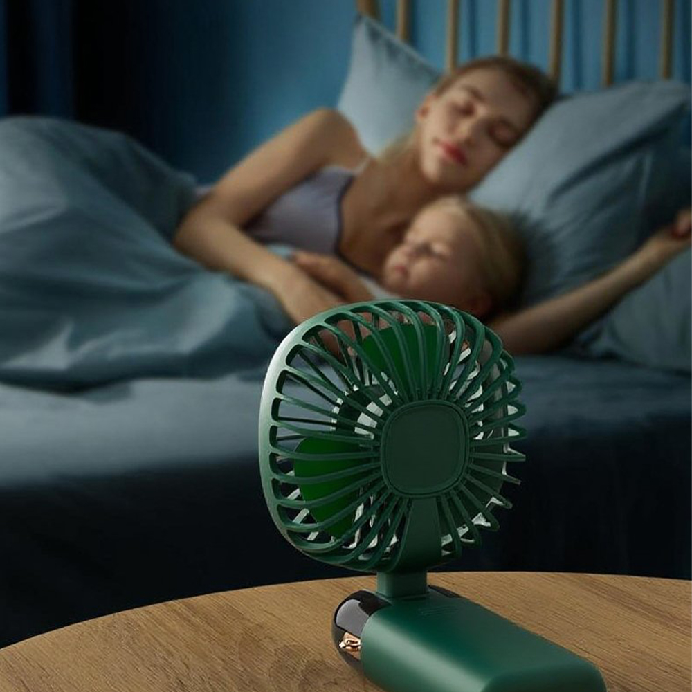 Ein grüner Handventilator in der Nähe schlafender Personen auf einem Bett in einem sanft beleuchteten Raum.