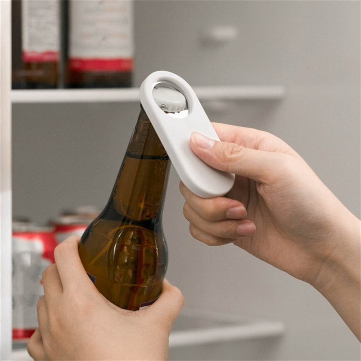 Eine Person öffnet eine Bierflasche mit einem weißen Flaschenöffner. Es ist vor einem Kühlschrank.