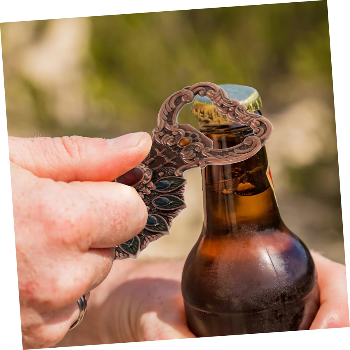 Hand öffnet eine Bierflasche mit einem bronzenen Flaschenöffner. Die Bierflasche ist braun und in der Sonne.