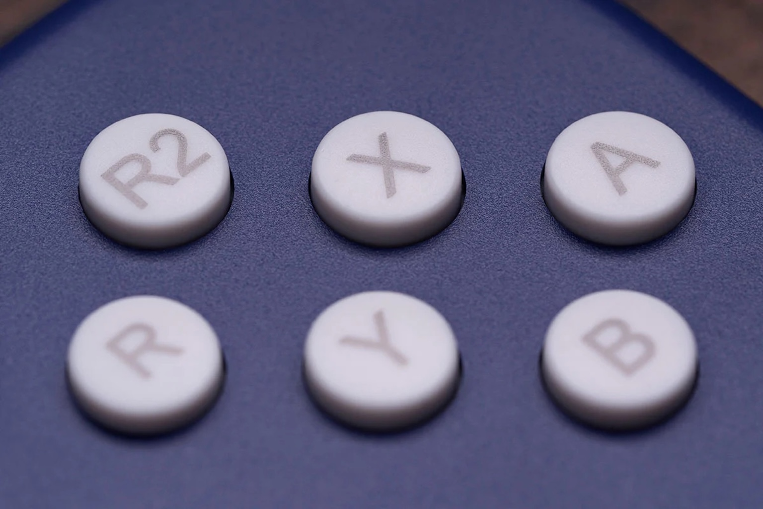 Primo piano dei pulsanti del controller: R2, X, A in alto; R, Y, B in basso. Tutti i pulsanti sono bianchi su una superficie blu.