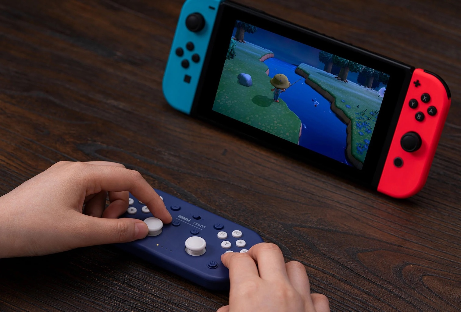 Mani che tengono un controller blu con pulsanti bianchi, accanto a una Nintendo Switch che mostra un gioco di Animal Crossing.