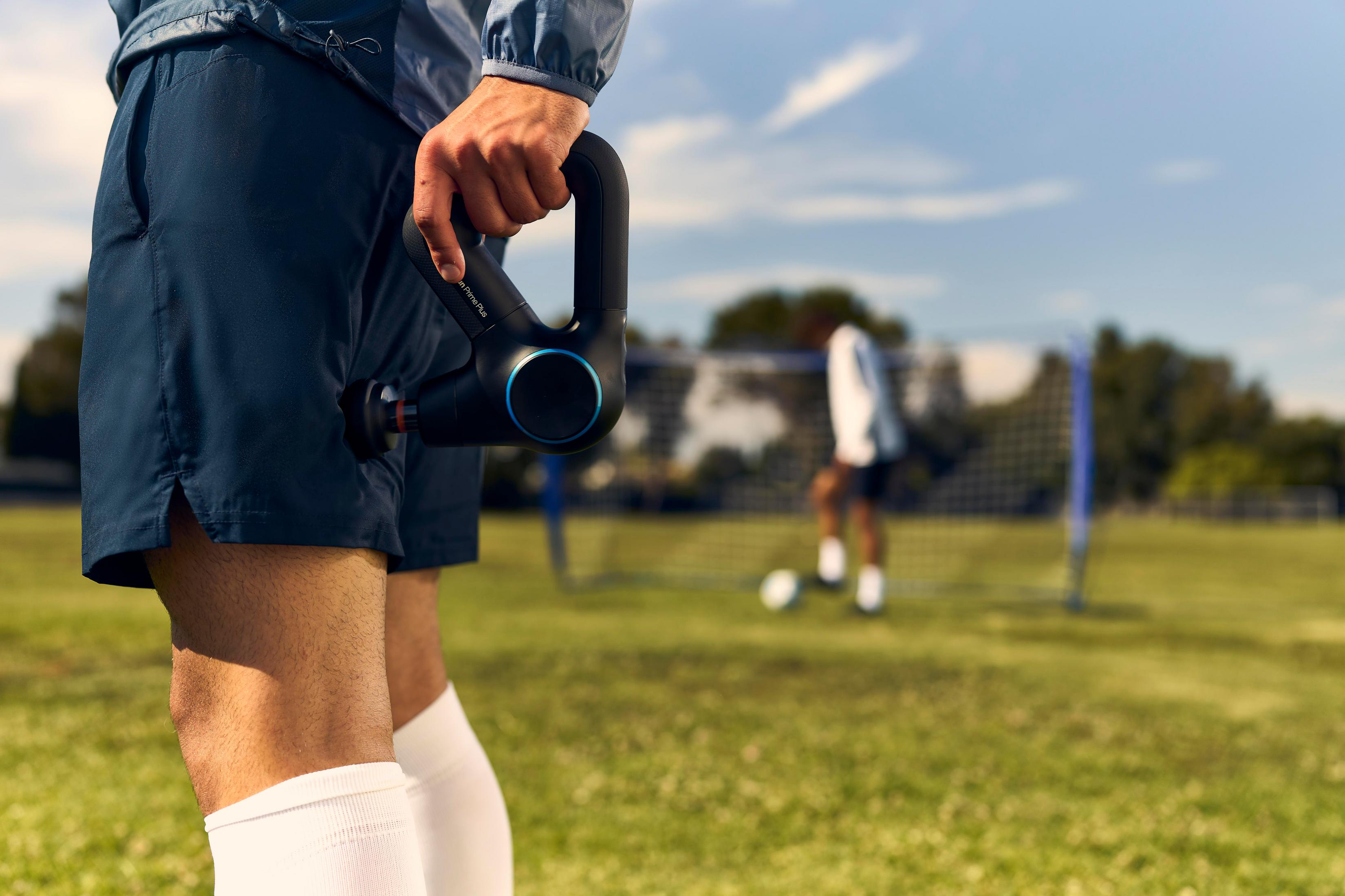 Osoba trzymająca czarny pistolet do masażu, szorty i białe skarpetki, na boisku z meczem piłki nożnej w tle.