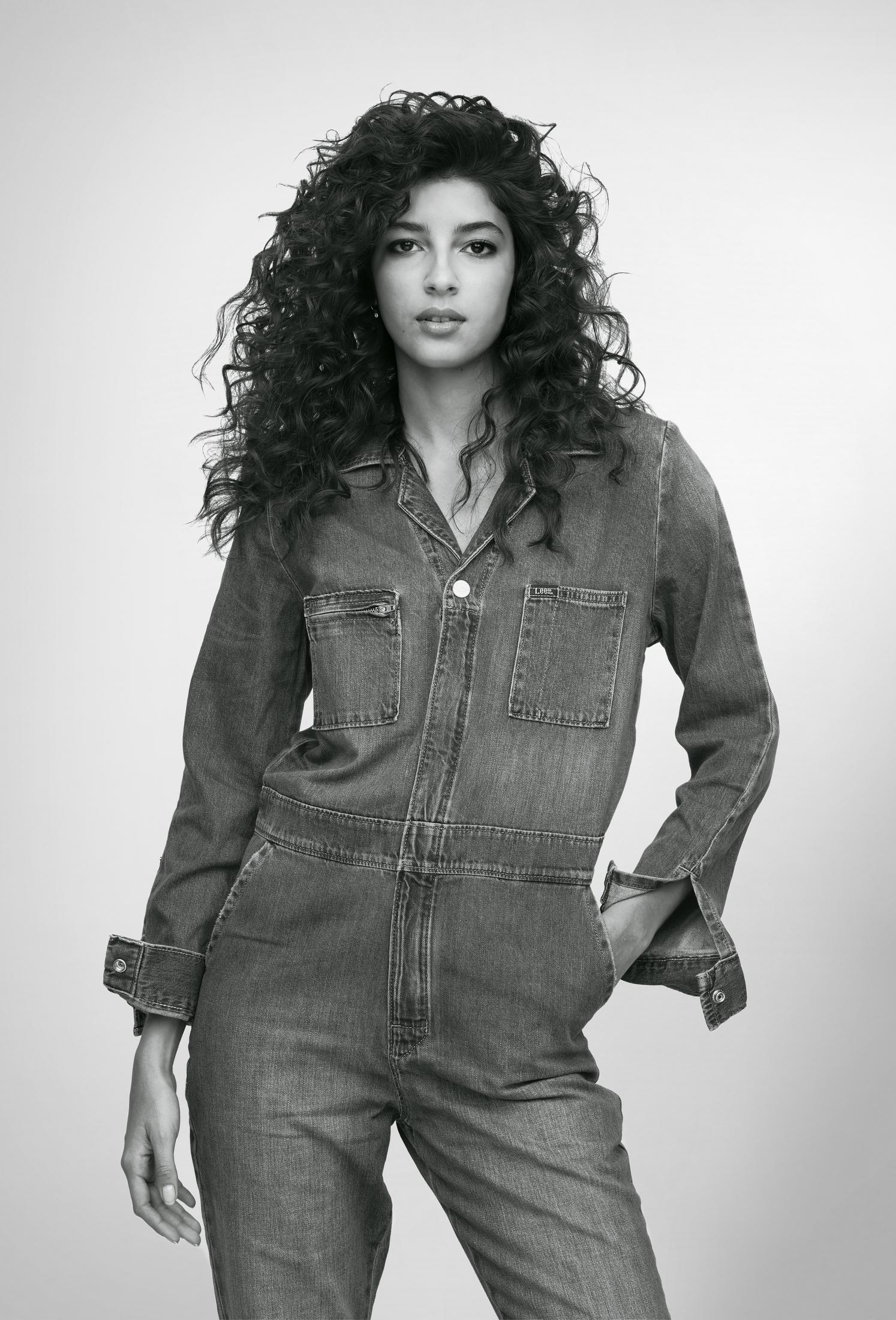 Ein Schwarz-Weiß-Foto einer Frau in einem Jeans-Overall. Sie hat lockiges Haar und blickt in die Kamera.