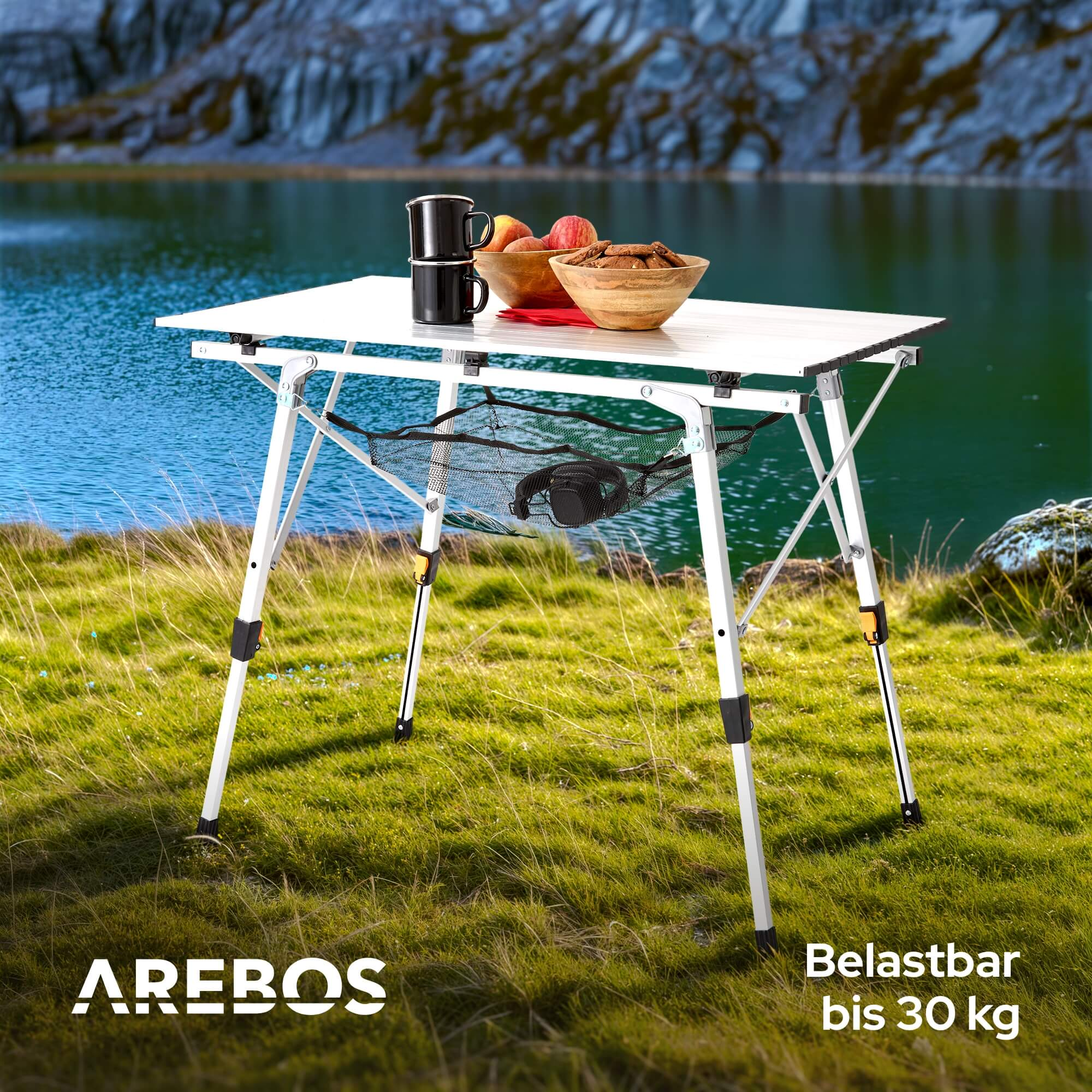 Ein Klapptisch auf Gras, mit Obst, Bechern und See im Hintergrund.