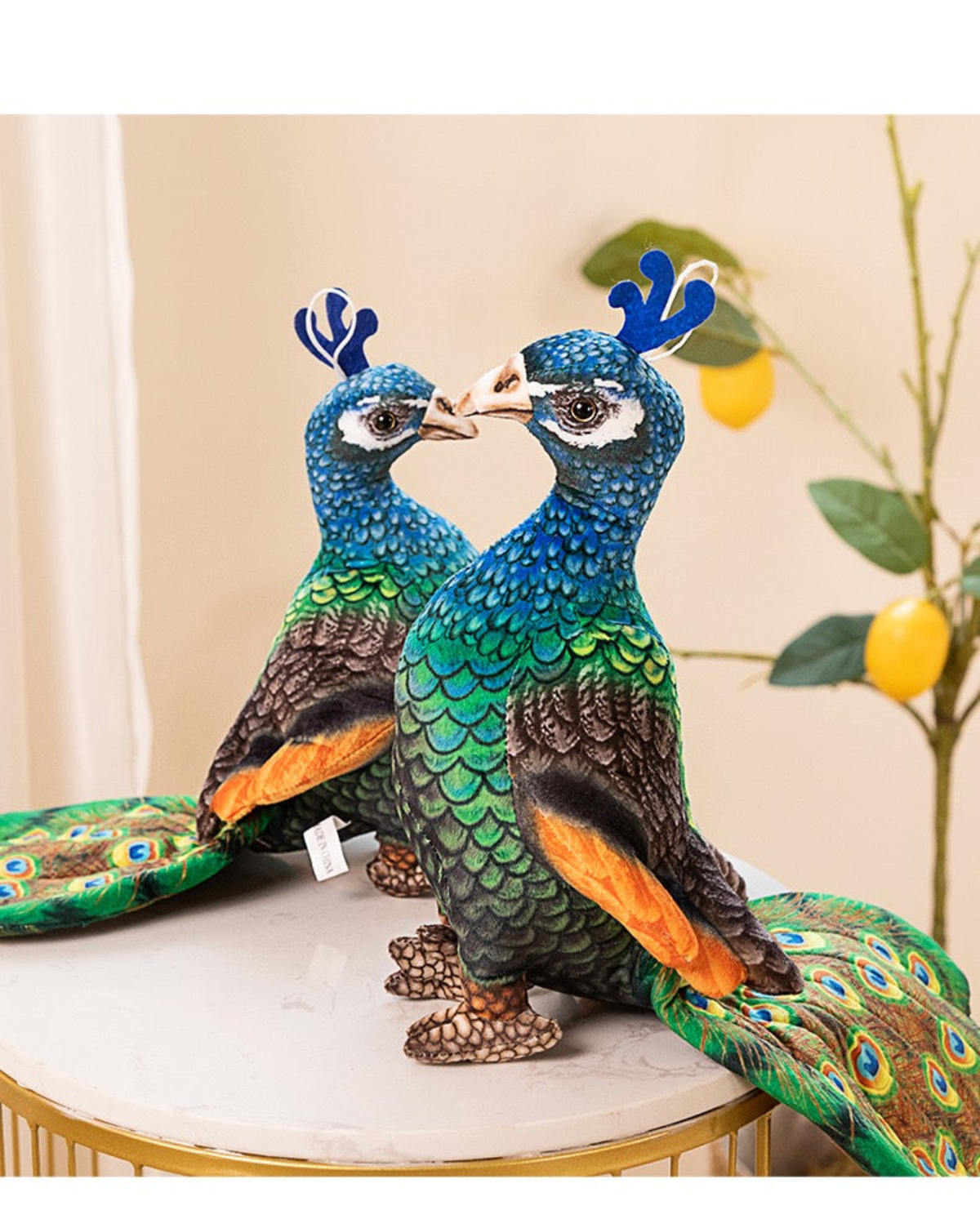 Zwei Pfau-Plüschtiere auf einer weißen Oberfläche. Sie sind blau, grün und braun, mit weißem Hintergrund und etwas Laub.