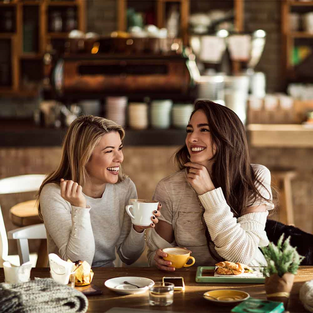 Zwei Frauen lächeln, halten Kaffeetassen in einem Café. Verschiedene Gegenstände auf dem Tisch, einschließlich Desserts.