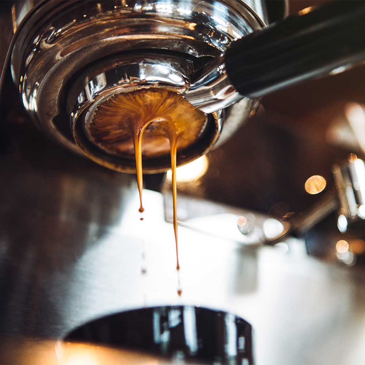 Nahaufnahme von Espresso, der aus einer Maschine fließt, zeigt dunkelbraune Flüssigkeit mit verschwommenen Hintergrundelementen.