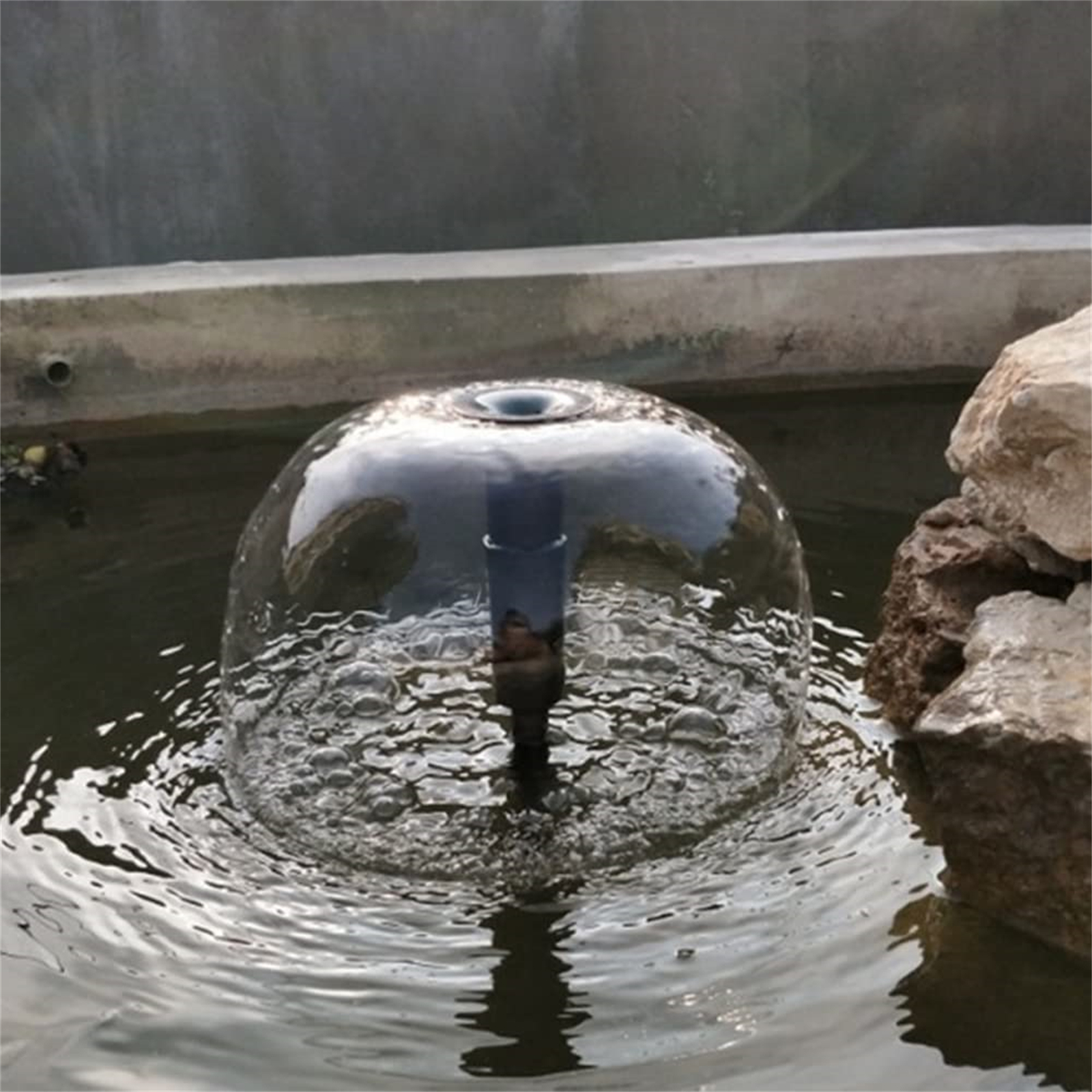 Ein Brunnen in einem Teich. Eine klare Wasserkugel sprüht nach oben. Das Wasser ist von Felsen und dunklem Wasser umgeben.